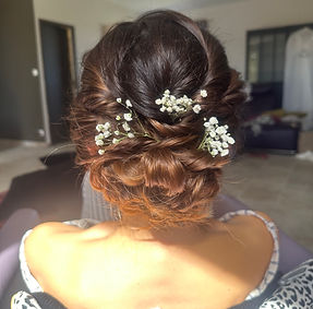 L’image montre une coiffure élégante vue de dos, avec des cheveux bruns soigneusement tressés et relevés en chignon. De petites fleurs blanches, probablement des gypsophiles, sont délicatement insérées dans la coiffure, ajoutant une touche de romantisme et de sophistication. La personne porte une robe à bretelles et une veste à motif léopard repose sur ses épaules. L’arrière-plan est flou, mais on peut distinguer une pièce intérieure avec des meubles et une porte. Cette image est intéressante pour illustrer des idées de coiffures de mariage ou d’événements spéciaux.