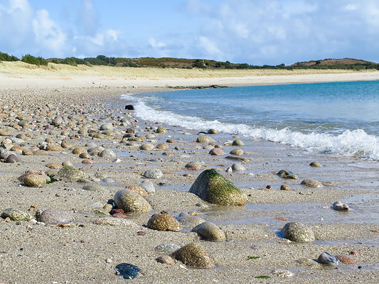 The shoreline of a sand beach with scattered cobbles and a light blue sea breaking, the sky is blue with the dunes visible in the background