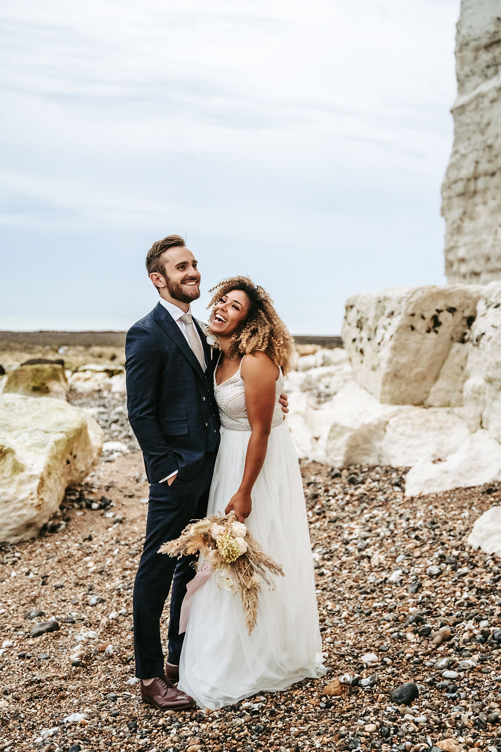 bride and groom laughing standing on the pebble beach in Seaford by the white cliffs at cuckmere haven, uk elopement photographer