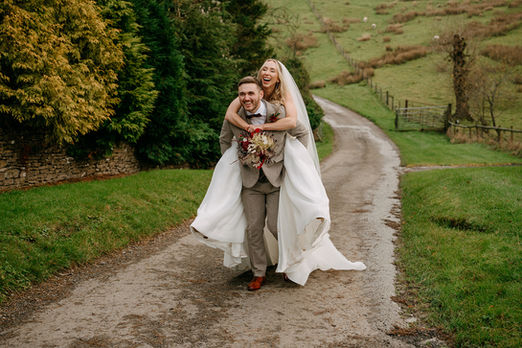 groom giving his bride a piggy back up a hill at Kingscote barn in the cotswolds