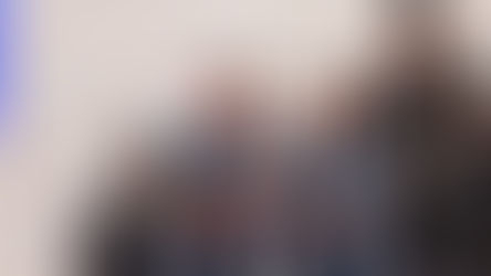 Winners in orange and blue suits hold a trophy on stage. Smiling man in cap, background with E1 and PIF logos, clapping people.