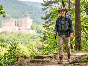 Ein älterer Mann mit grauem Bart, Wanderhut, kariertem Hemd und großem Wanderrucksack geht lächelnd einen wurzeligen Waldpfad entlang. Er nutzt einen Wanderstock. Im Hintergrund thront auf einem bewaldeten Hügel das Hambacher Schloss mit Deutschlandfahne unter einem hellen Himmel.