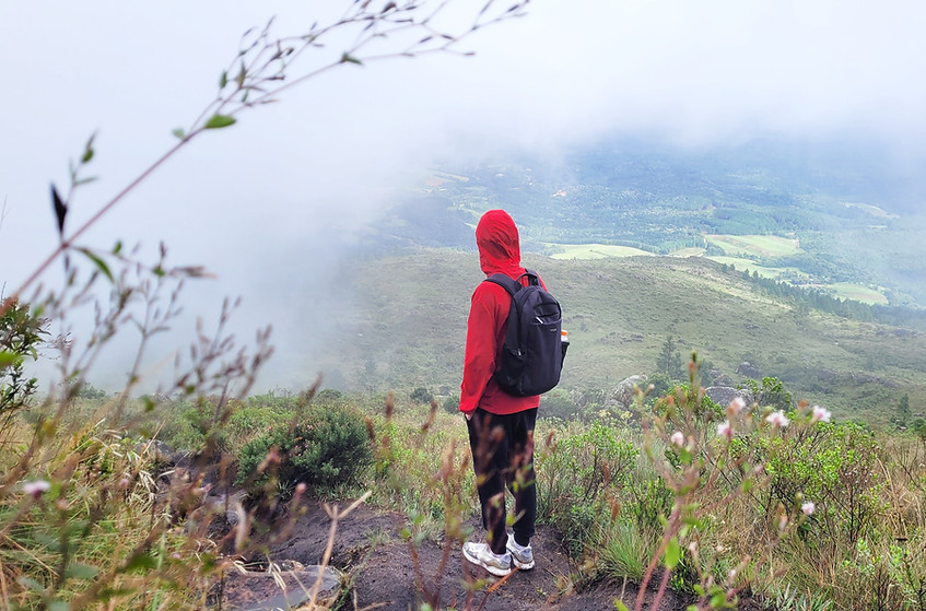 Jovem vestindo jaqueta vermelha está em pé, em meio a natureza, olhando em direção as montanhas e nuvens à sua frente.