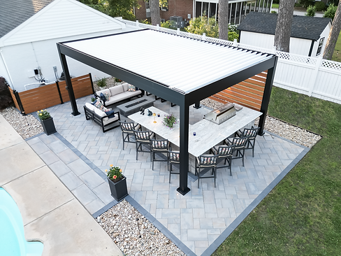 Overhead view of a sleek, modern outdoor living area featuring a black Azenco louvered pergola. Beneath the pergola is a large outdoor kitchen with bar seating, complemented by a cozy lounge area with sofas and a fire feature. The space is framed by elegant pavers and landscaped accents