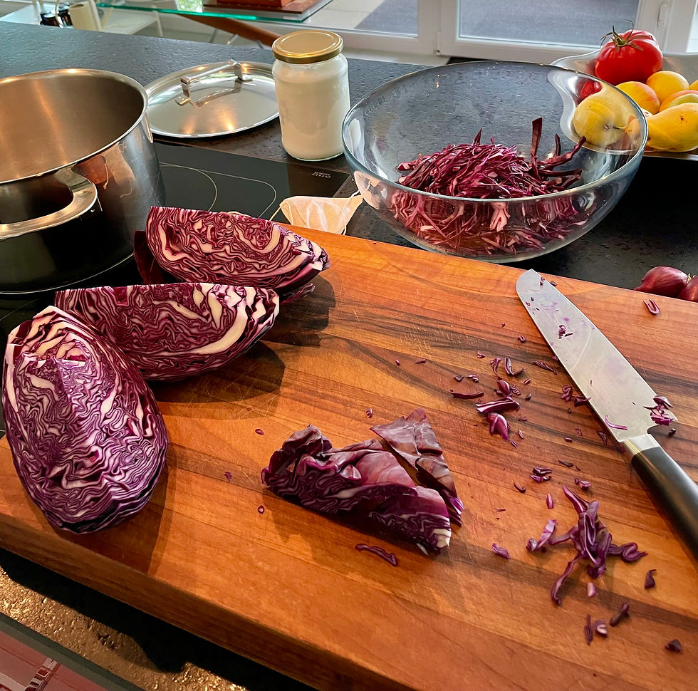 Red cabbage being sliced on a wooden cutting board in a bright kitchen, with knife, pot, bowl of shreds, and fruit nearby
