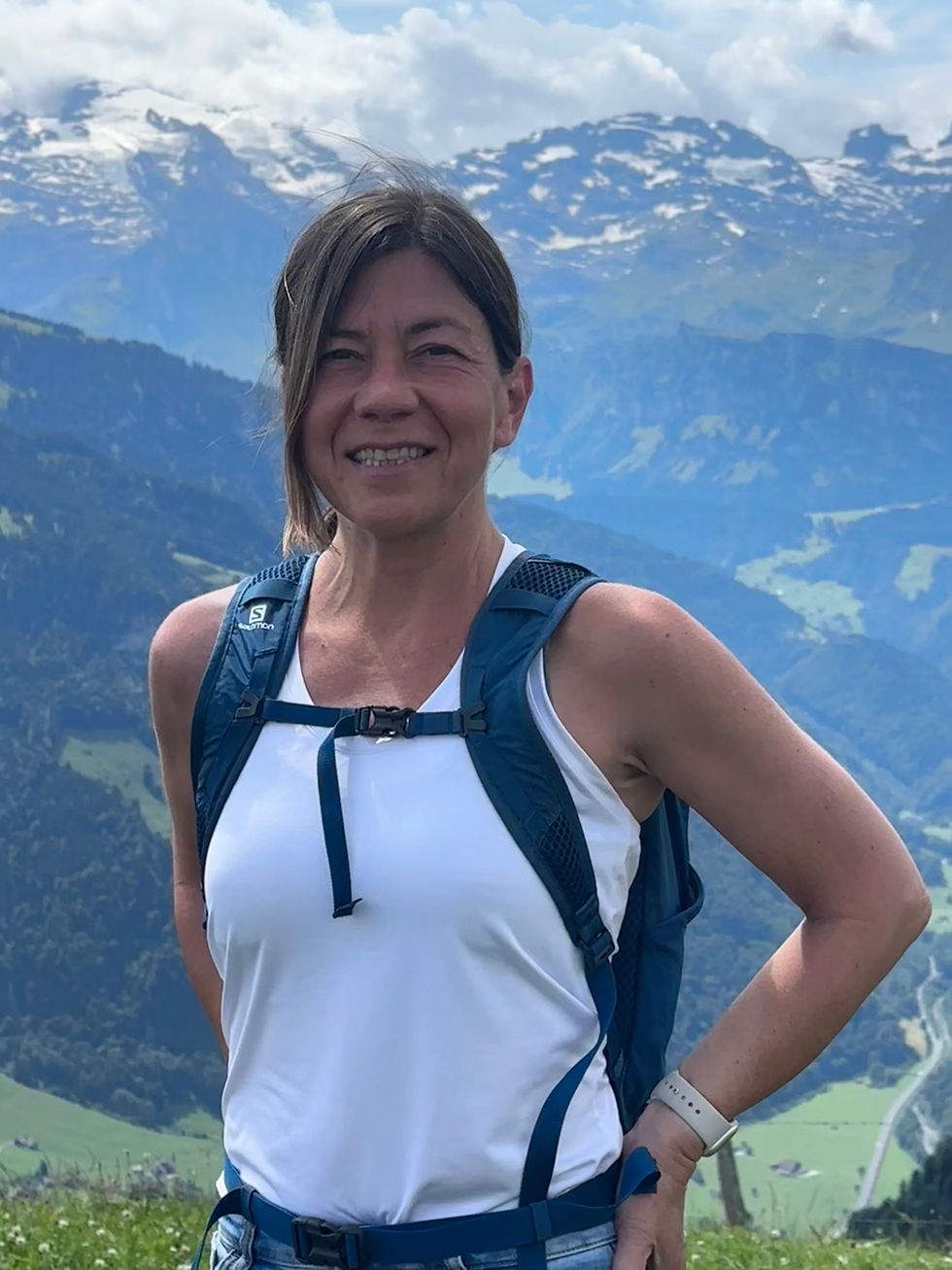Smiling woman with a blue backpack stands against a scenic mountain backdrop on a clear day, wearing a white tank top. Snow-capped peaks visible.