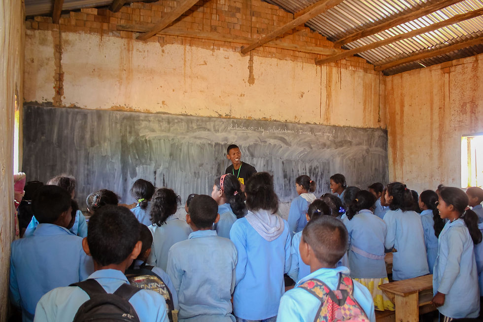 A male Projet Jeune Leader educator stands at the front of a large run-down classroom in rural Madagascar, leading the room of students in an activity.