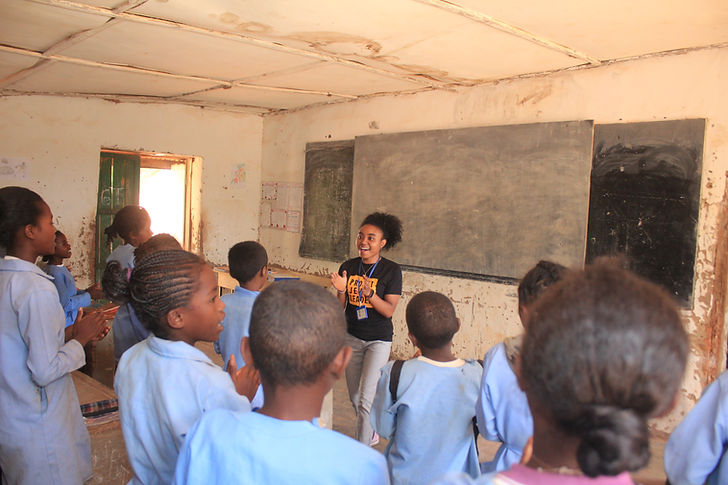 This is a photo of a group of children in blue uniforms in a classroom standing and clapping. The classroom has a blackboard in the background. At the front of the classroom is a female Projet Jeune Leader Educator clapping and smiling. The walls of the classroom are peeling and the ceiling is falling apart.