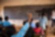 A photo of a middle school student in Madagascar raising her hand, one finger in the air. The background faintly shows a classroom scene with other seated students.