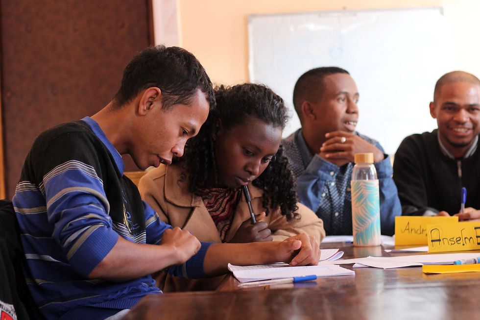 One young Malagasy man and woman sit next to each other intently looking at a piece of paper. In the background, two young men also sit at the table smiling and talking with each other.
