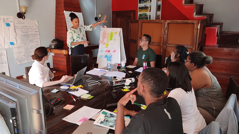 A group of people sit around a large table and look at a woman speaking and gesturing to a flipchart paper covered in post-it notes.