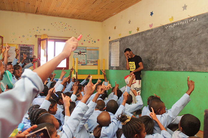 A male Projet Jeune Leader educator is laughing while holding his head in his hand. He is standing in front of a blackboard in a classroom facing rows of students sitting on the ground. The students are laughing and energetically raising their hands.