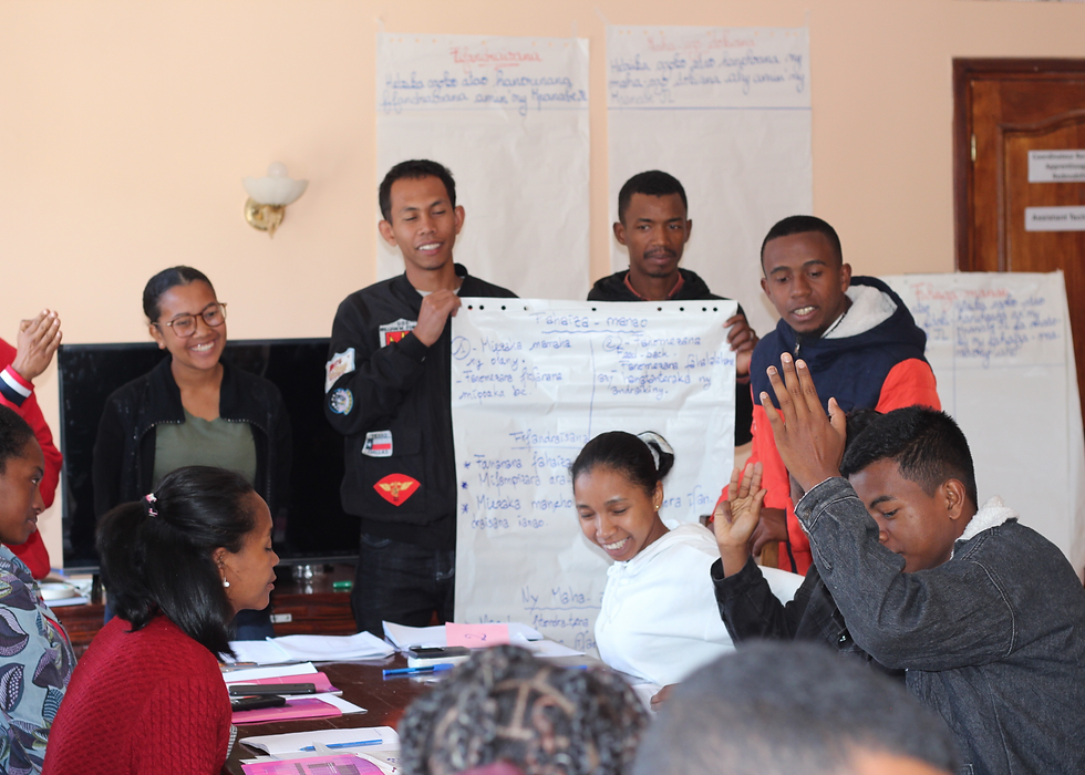 Around ten of Projet Jeune Leader's new technical coordinators sit and stand around a table. Some are holding a large flipchart paper; others are smiling and clapping.