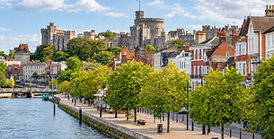 Windsor Castle towers above a riverside promenade lined with trees and historic buildings.