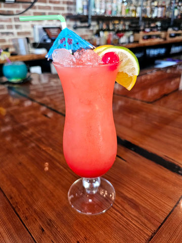 A tall, curvy glass of red Crab Trap punch, garnished with a blue cocktail umbrella, oranges, limes, and cherries