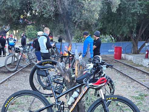 Mountain bikes parked on tree stumps near railway tracks, with cyclists gathered around a fire pit at Calitzdorp Station in the Klein Karoo.

