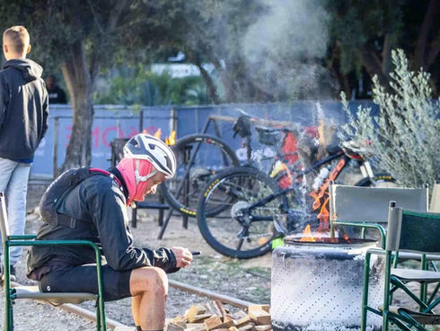 Cyclist in helmet and gear sitting next to a fire pit on railway tracks at Calitzdorp Station, with bicycles and outdoor seating in the background.