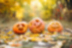 Three carved pumpkins with various faces sit on a leaf-covered path. The background is a vibrant, blurry autumn scene in warm colors.