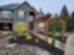 A gray stone house with a pergola and landscaped garden. Vibrant plants, a stone path, and a parked car are in the driveway. Overcast sky. Beautiful new bark mulch in the landscaping.