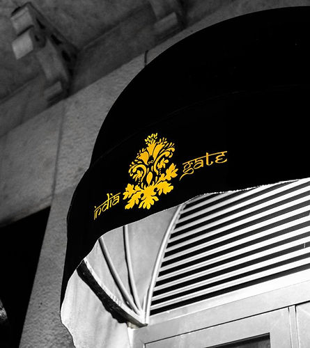Black-and-white exterior view of India Gate Indian restaurant with a black awning featuring the gold India Gate logo and emblem above the entrance.