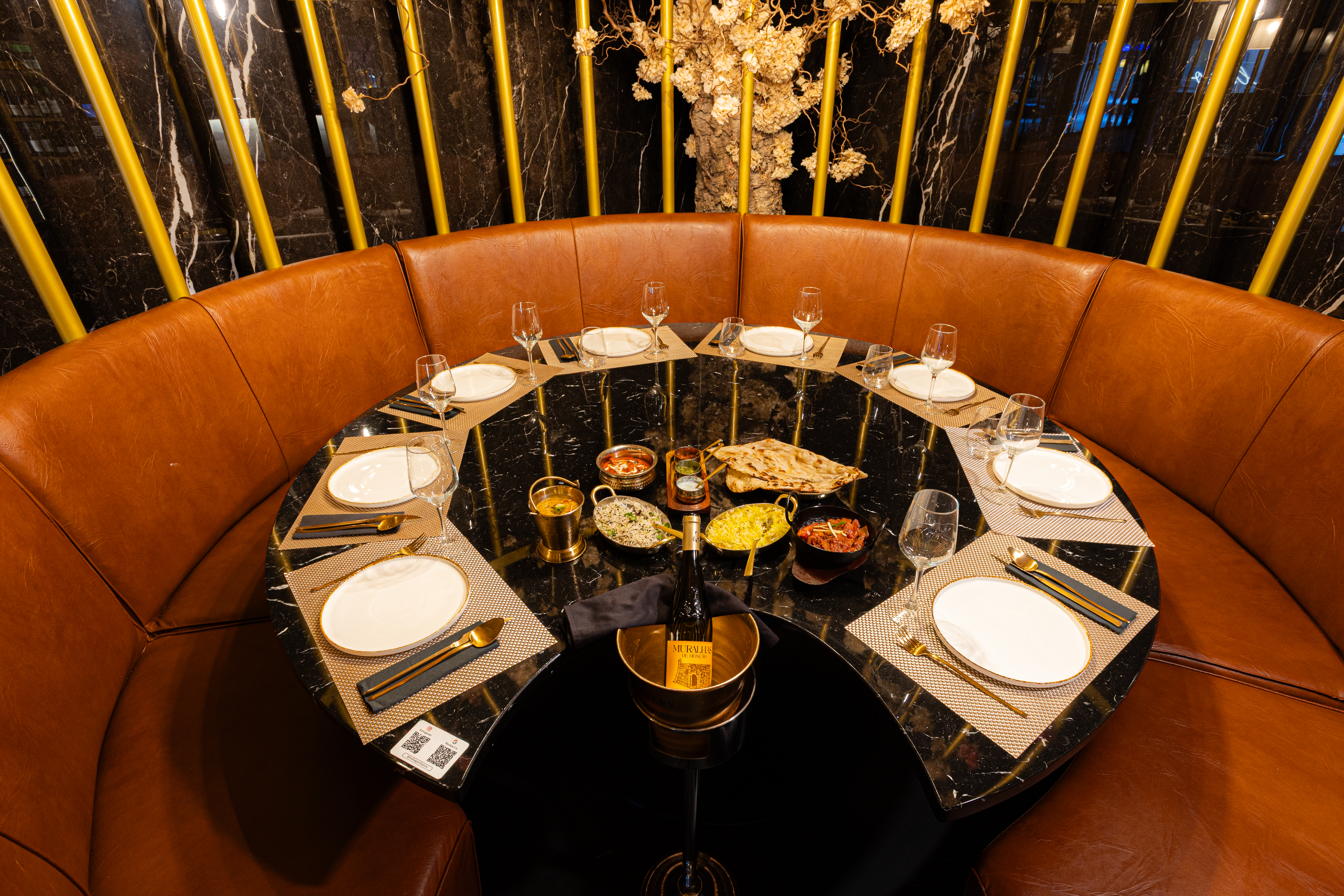 A luxury private dining setup at India Gate Santos in Lisbon, featuring the restaurant’s iconic golden floral dome, a round black marble table beautifully set for ten guests with elegant plates, gold cutlery, crystal glassware, and premium wine. Authentic Indian dishes including curries, biryani, naan, and sides are arranged at the center, showcasing the fine-dining catering experience and premium ambience India Gate offers for private events, corporate dinners, group celebrations, and high-end Indian dining in Lisbon.