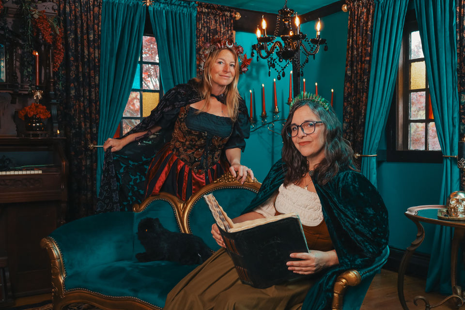 A woman in a tan and brown peasant-style dress and teal cape sits on a velvet teal chaise lounge with gold trim, in a room with teal walls and a mixture of teal velvet and black floral velvet curtains. Another woman in a teal and red dress with a black cape stands behind the chaise lounge; each woman is wearing a flower crown. There is a grand piano with a red velvet chair to the left, and a black chandelier hanging in the back right corner.