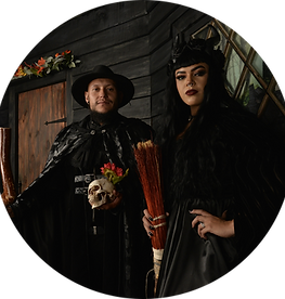 A man and a woman standing in front of a set of a dark colonial-home-style backdrop; the man is in a cape and hat holding a skull with roses, and the woman is in a black cape and black headpiece with horns, holding a broom