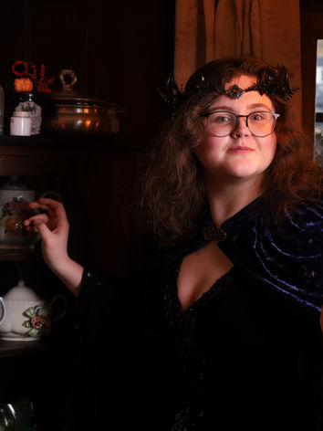 A woman with shoulder length red hair, wearing a black floral headpiece, a black dress, and a purple cape, stands in front of dark wooden shelves adorned with bottles, flowers, and teapots, ready to make a selection, but facing the camera with a smile