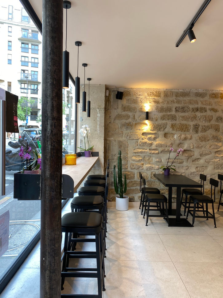 Immergez-vous dans l'atmosphère unique du restaurant, où l'élégance et l'authenticité se rencontrent dans chaque détail architectural. En entrant, vos yeux sont immédiatement attirés par le bar en acier et bois suspendu, créant un contraste saisissant avec la vitrine du restaurant. Cette conception audacieuse ajoute une touche de modernité tout en conservant une ambiance chaleureuse et accueillante.

Le mur en pierre, qui se marie parfaitement avec les tables en bois rustique, évoque un charme intemporel et une sensation de stabilité. Chaque table, soigneusement disposée sur le carrelage gris du sol, invite les convives à s'attarder et à profiter de l'atmosphère conviviale du lieu.

Dans cet espace, l'acier, le bois, la pierre et le carrelage se fondent harmonieusement pour créer une expérience sensorielle unique, où chaque élément architectural contribue à l'ambiance générale du restaurant. C'est un véritable havre de convivialité où la qualité de l'accueil et la délicatesse du design se rejoignent pour offrir une expérience culinaire inoubliable.
