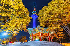Seoul Namsan Tower (N Seoul Tower) at night, glowing blue against bright yellow autumn trees and traditional pavilion