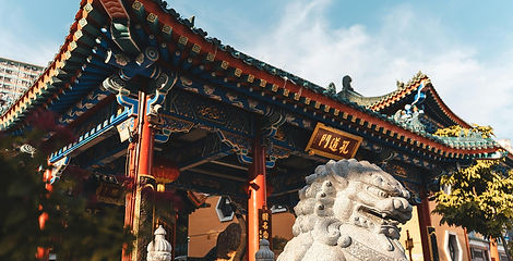A carved stone guardian lion stands in front of the ornate traditional multi-tiered roof and red entrance gate at Wong Tai Sin Temple in Hong Kong.