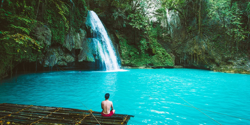 A man sits on a bamboo raft in a vibrant turquoise pool beneath a cascading waterfall surrounded by lush green jungle cliffs at Kawasan Falls in Cebu.