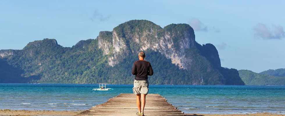 A traveler walks along a rustic wooden pier toward the stunning turquoise ocean in Palawan, featuring a limestone mountain and a traditional Filipino bangka.