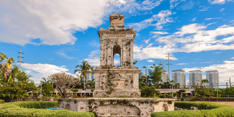 A historic stone obelisk monument dedicated to Magellan stands in a manicured garden park against a blue sky in Mactan, Cebu.