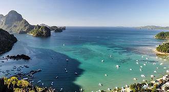 A stunning high-angle view of El Nido town and Bacuit Bay from Taraw Cliff, showing numerous boats anchored in turquoise waters surrounded by limestone karsts.