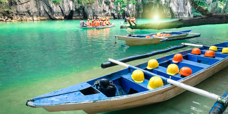 Tourists in orange life vests sit on a wooden paddle boat on clear turquoise water near the limestone cave entrance of the Puerto Princesa Underground River.