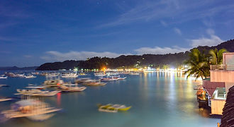 The peaceful bay of El Nido town at night, showing lit-up beachfront establishments and traditional outrigger boats anchored on the calm water under a dark sky.