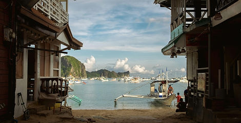 Outrigger boats sit on shallow shoreline waters viewed through a wooden beachfront street in El Nido town, Palawan, Philippines.