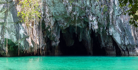 Hanging vines drape over the dark limestone entrance of the Puerto Princesa Underground River cave above clear turquoise water.