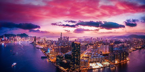 A high-angle panoramic aerial view of Victoria Harbour and the Hong Kong city skyline glowing with lights under a vibrant pink and purple twilight sky.
