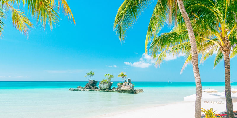 Shallow turquoise sea waters surround the rocky islet of Willy's Rock beneath swaying palm trees along powdery White Beach in Boracay, Philippines.