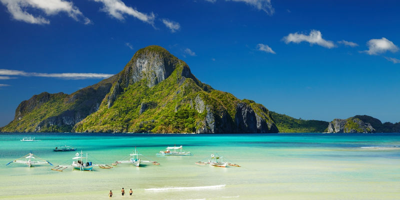 Outrigger boats float on shallow clear turquoise water facing the lush green mountain peak of Cadlao Island under a bright blue sky in El Nido, Philippines.