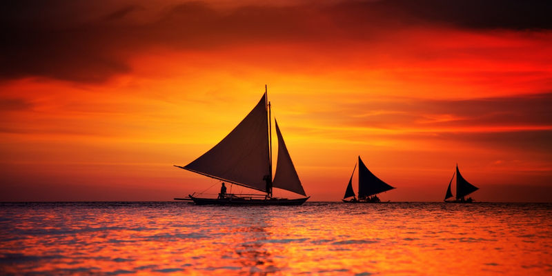 Silhouetted paraw sailboats sail across calm ocean waters beneath a dramatic orange and red sunset on White Beach in Boracay, Philippines.
