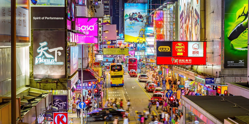 Glowing neon billboards and brightly lit store signs overhang a busy street filled with double-decker buses and pedestrians in Mong Kok, Hong Kong.
