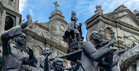 Detailed dark bronze statues depicting historical figures and religious icons stand in front of stone church reliefs at the Heritage of Cebu Monument.