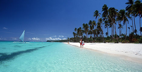 A white and blue traditional outrigger boat rests on shallow clear turquoise water along a white sand beach under a bright blue sky in Boracay, Philippines.
