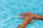 Two wet hands with pink nails and shell bracelets resting in bright blue pool water - summer nail care tips from Zen Nail Spa