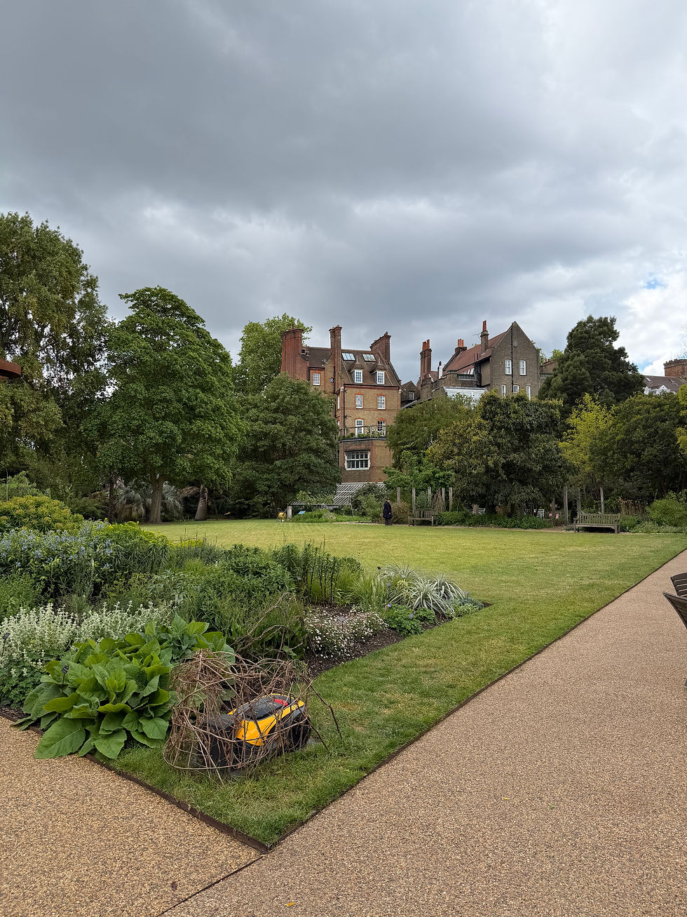 Formal garden with lawns, trees, and paths under cloudy sky, backed by brick houses and a lone distant figure
