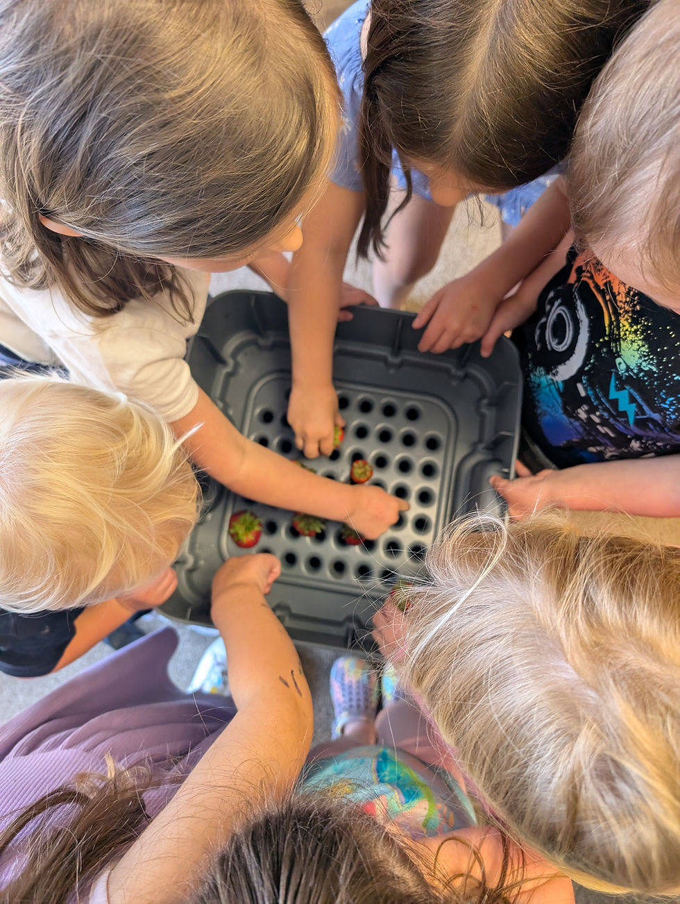 Children gather around a black tray filled with strawberries, reaching in eagerly. The setting is bright and playful, capturing curiosity.