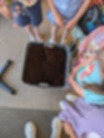 Children in colorful outfits stand around a bin of soil on a carpeted floor. One child points at the soil, indicating interest.